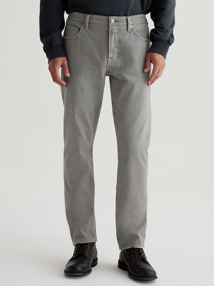 A person wearing AG Jeans Everett in 7 Years Sulfur Ironwood paired with a dark long-sleeve shirt and black lace-up boots stands against a plain light background.