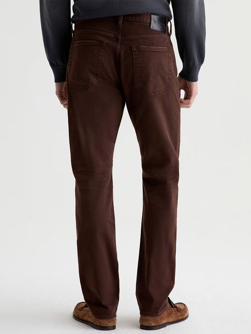 A person stands facing away, wearing AG Jeans Everett in 7 Years Sulfur Wood Brown, a dark sweater, and brown shoes, against a plain light background.