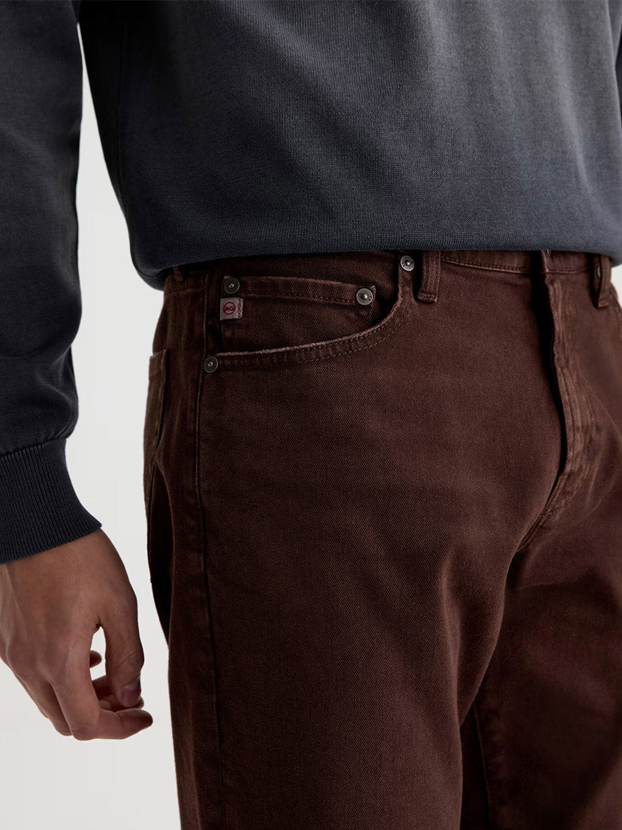A person wearing a dark gray sweatshirt and AG Jeans Everett in 7 Years Sulfur Wood Brown, shown from mid-torso to thigh with focus on the pocket area.