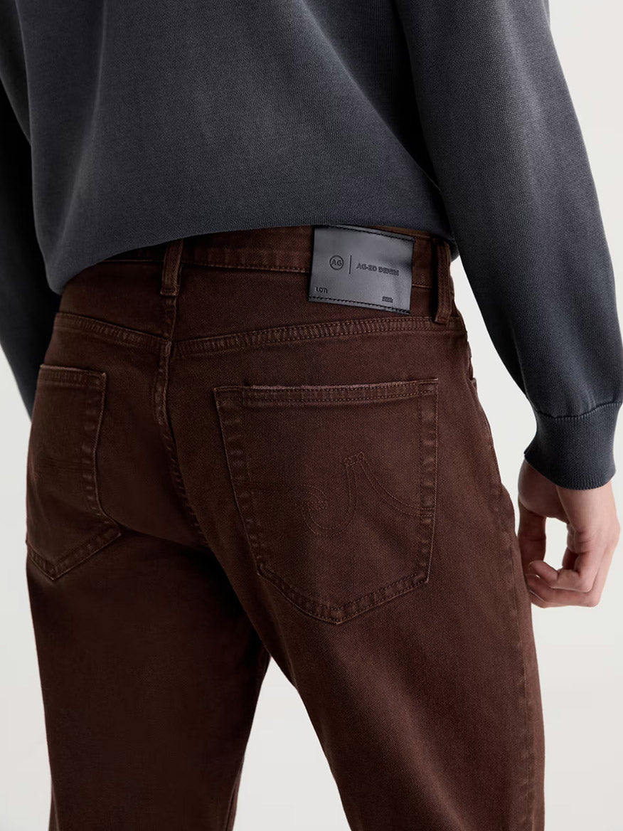 Person wearing AG Jeans Everett in 7 Years Sulfur Wood Brown, styled with a dark long-sleeve top and shown from the back, highlighting the visible black label on the waistband. Crafted from AG Cloud Soft Denim.
