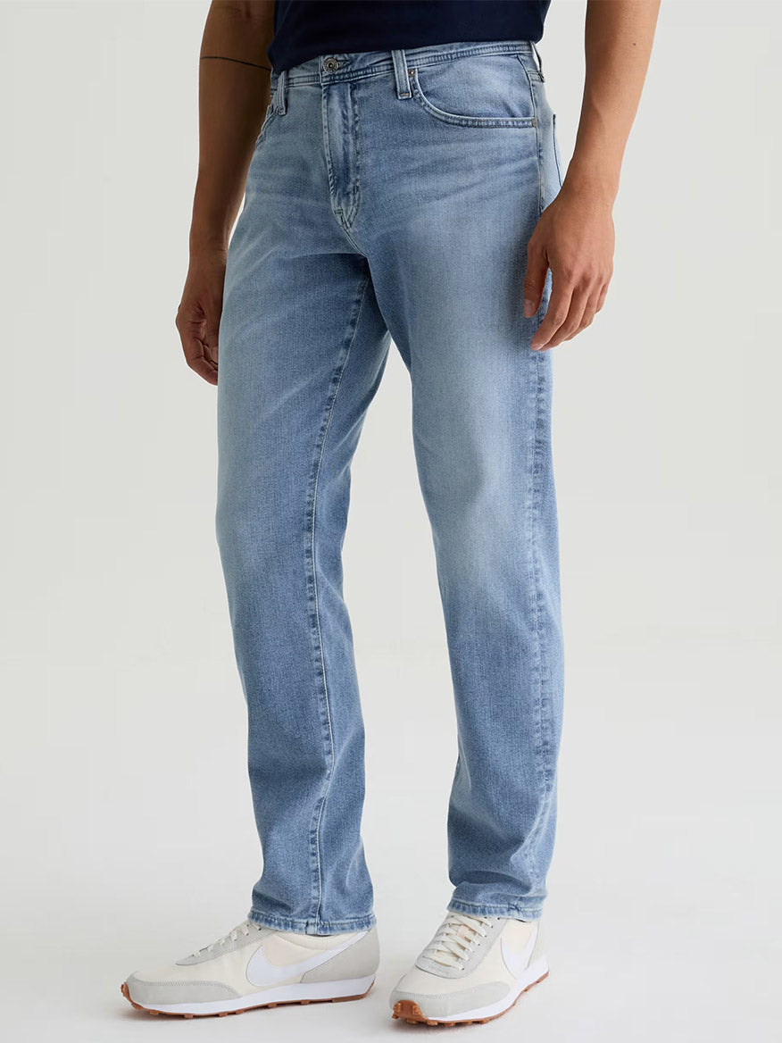 A person wearing AG Jeans Everett in Saltillo, a navy shirt, and white sneakers stands against a plain light background.