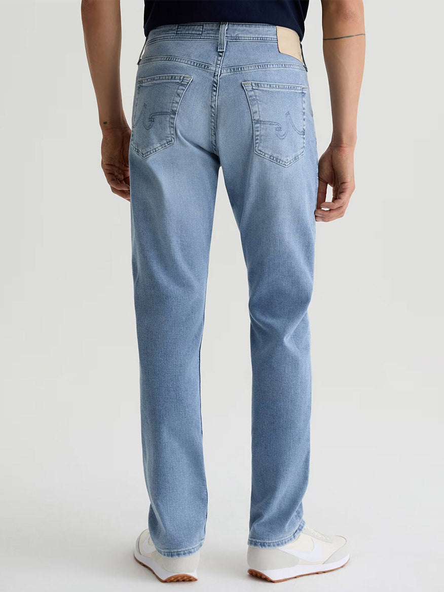 A person, shown from the waist down and facing away, wears AG Jeans Everett in Saltillo (a light indigo wash men’s slim straight jean), paired with white sneakers and a dark shirt against a plain background.