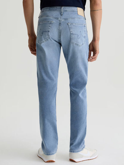 A person, shown from the waist down and facing away, wears AG Jeans Everett in Saltillo (a light indigo wash men’s slim straight jean), paired with white sneakers and a dark shirt against a plain background.