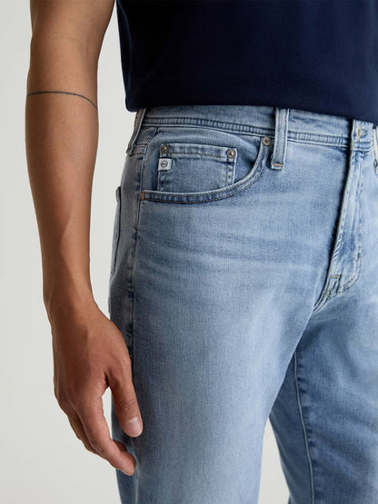 A person wearing AG Jeans Everett in Saltillo, slim straight jeans in light blue Cloud Soft Denim, paired with a dark navy shirt and revealing part of an arm with a thin, straight tattoo.