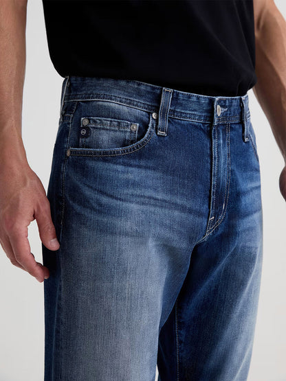 A person wearing AG Jeans Everett in Upton, a men’s slim straight jean, paired with a black t-shirt, shown from waist to mid-thighs with one hand visible at their side.