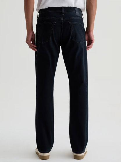Person standing facing away, wearing AG Jeans Everett 360° Jean in Vintage Ink, a white shirt tucked in, and white sneakers against a plain background.