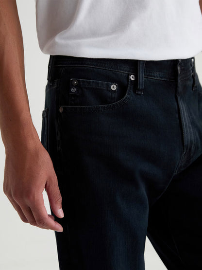 A person is shown from waist to mid-thigh wearing a white t-shirt and AG Jeans Everett 360° Jean in Vintage Ink, with one hand resting by their side.