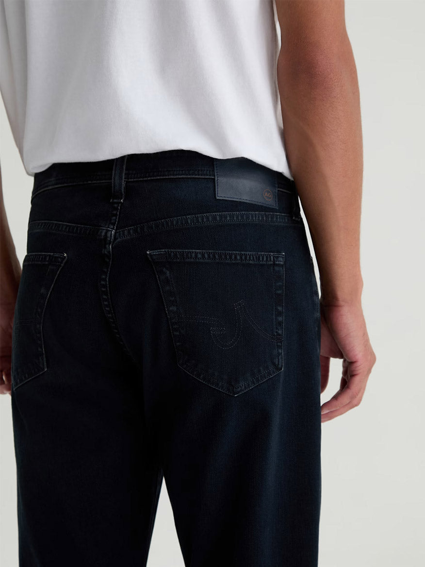 A person shown from the waist down, with their back to the camera, wears a white shirt and AG Jeans Everett 360° Jean in Vintage Ink.