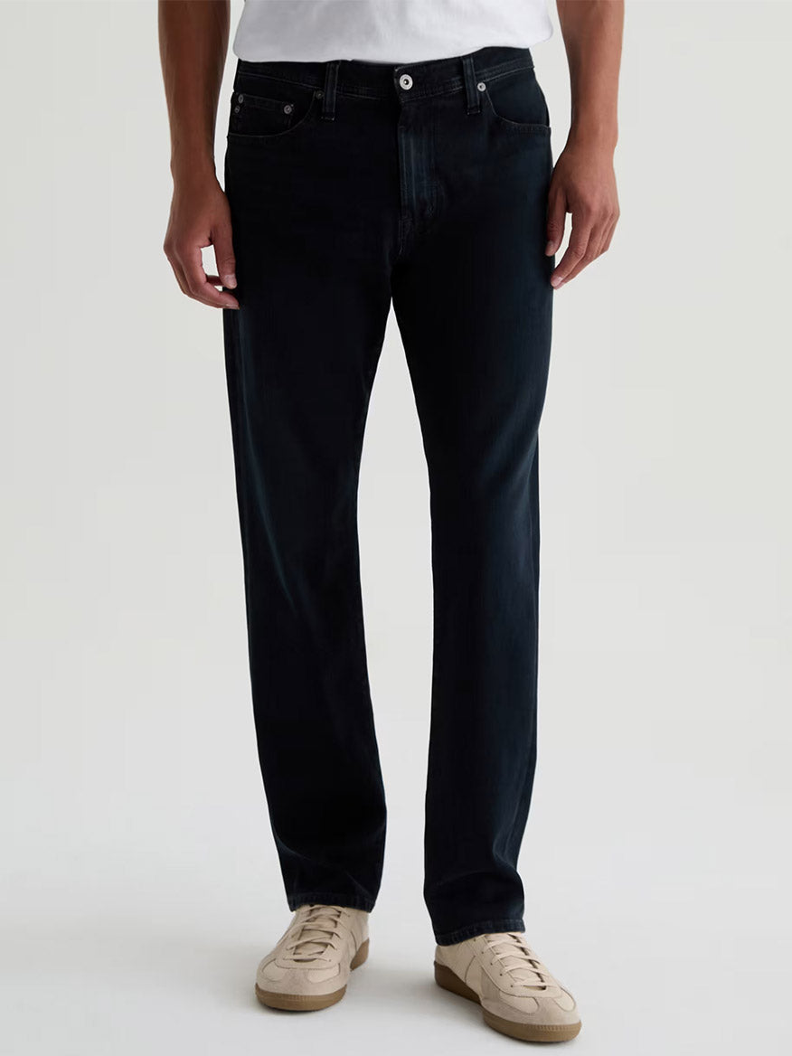 A person wears a white shirt, AG Jeans Everett 360° Jean in Vintage Ink, and beige sneakers, standing against a plain light background.