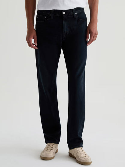 A person wears a white shirt, AG Jeans Everett 360° Jean in Vintage Ink, and beige sneakers, standing against a plain light background.