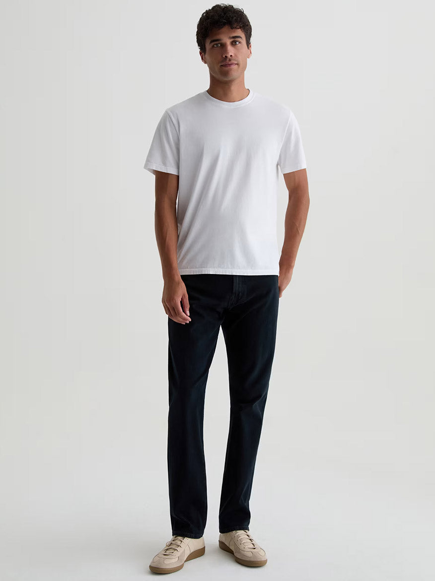A man stands against a plain background wearing a white short-sleeve t-shirt, beige sneakers, and AG Jeans Everett 360° Jean in Vintage Ink—slim straight, dark blue stretch denim.