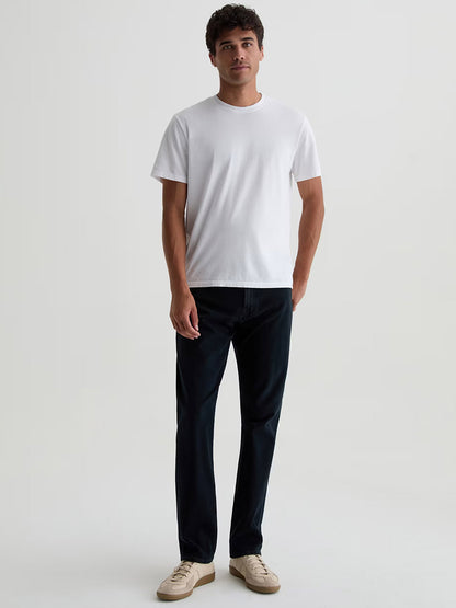 A man stands against a plain background wearing a white short-sleeve t-shirt, beige sneakers, and AG Jeans Everett 360° Jean in Vintage Ink—slim straight, dark blue stretch denim.