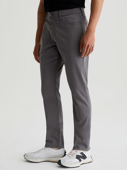 A person wearing AG Jeans Everett Sueded in Folkestone Grey, a black shirt, and white New Balance sneakers stands against a plain light background.