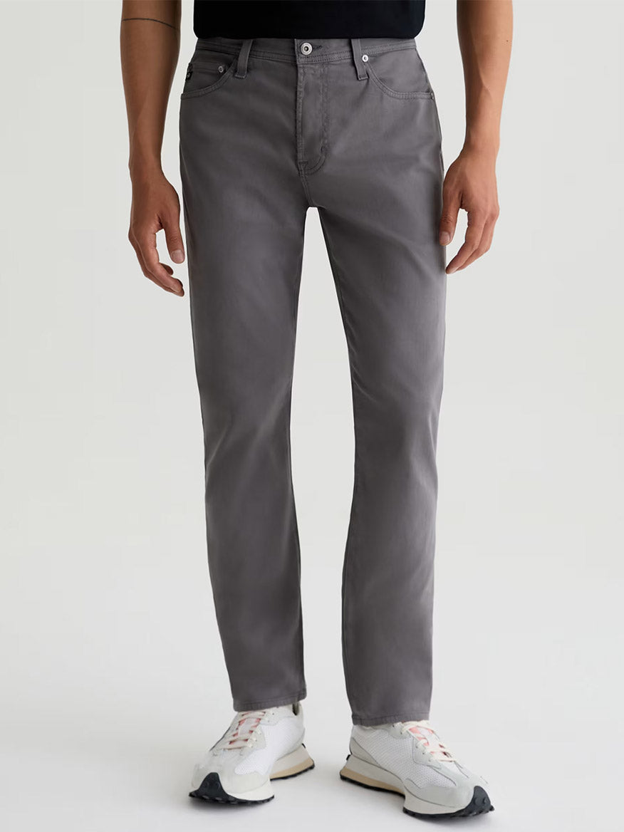 Someone wearing AG Jeans Everett Sueded in Folkestone Grey, a black shirt, and white sneakers stands against a plain light background.