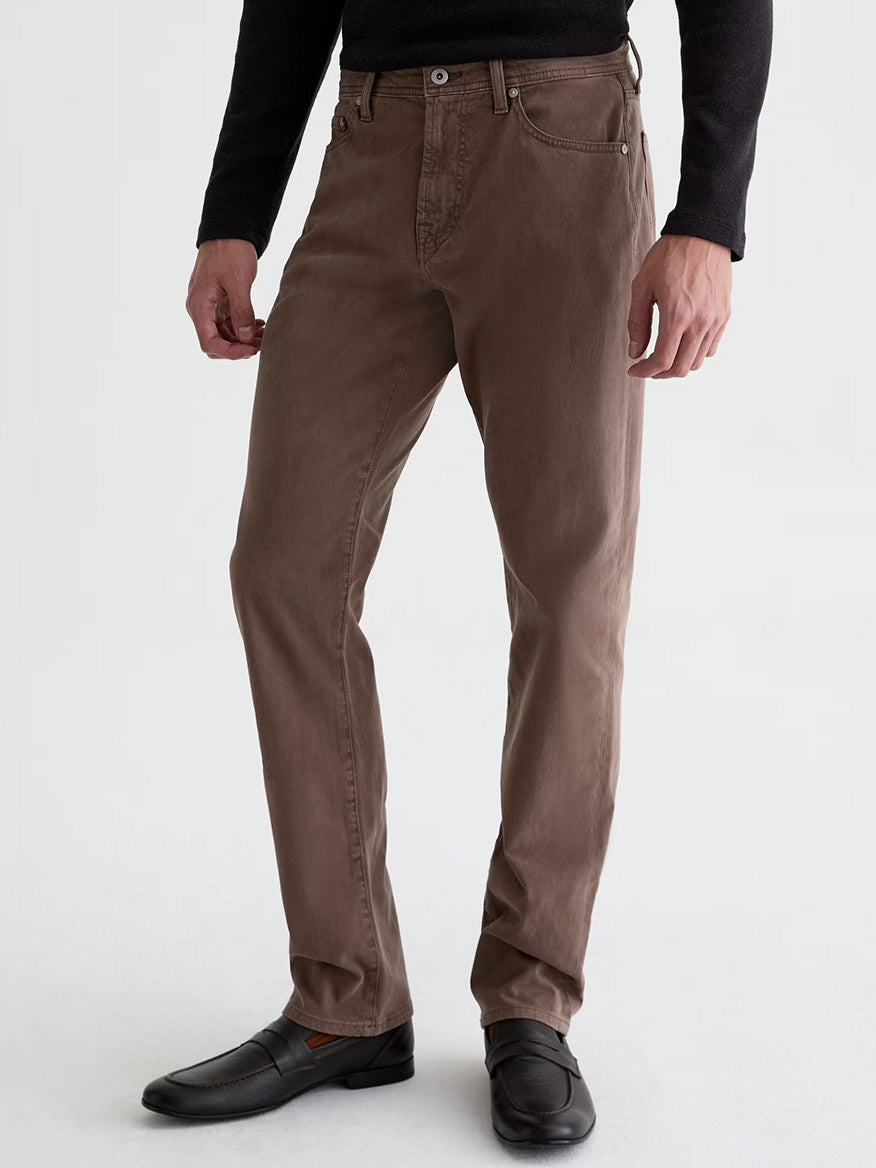 A man wears the AG Jeans Everett Sueded in Sulfur Taupe Grey, paired with a black long-sleeve shirt and black dress shoes, standing against a plain light background for a subtle vintage look.