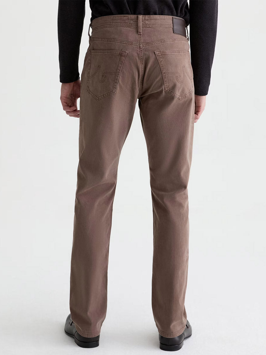 A person stands facing away, wearing the AG Jeans Everett Sueded in Sulfur Taupe Grey—a slim straight jean in stretch sateen—paired with a black long-sleeve top and black shoes, set against a plain light background.