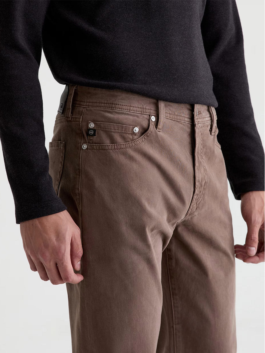 Wearing the AG Jeans Everett Sueded in Sulfur Taupe Grey—a pair of brown slim straight jeans in a sueded stretch sateen finish—paired with a long-sleeve black shirt, the person stands with arms relaxed at their sides, shown from mid-torso to mid-thigh. The jeans offer a vintage look that complements the classic style.