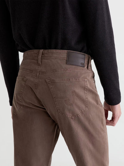 Model shown from the back wearing AG Jeans Everett Sueded in Sulfur Taupe Grey paired with a black long-sleeve shirt. Crafted from comfortable sueded stretch sateen, these slim straight jeans feature stitched back pockets and a dark leather patch on the waistband, adding a subtle vintage touch.