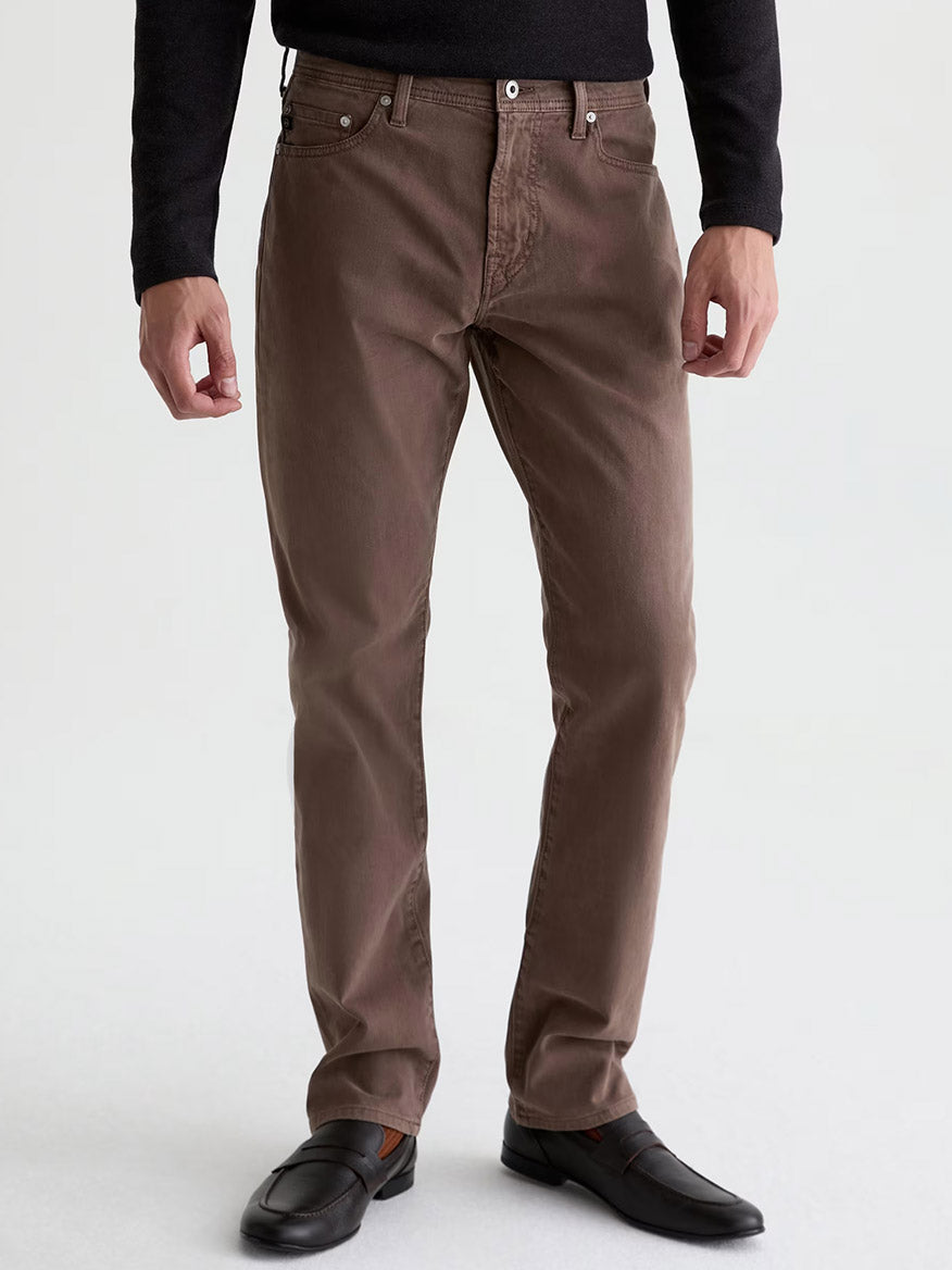 Wearing the AG Jeans Everett Sueded in Sulfur Taupe Grey—brown slim straight jeans made from sueded stretch sateen—a person pairs them with a black long-sleeve shirt and black loafers, standing against a plain background for a subtle vintage look.