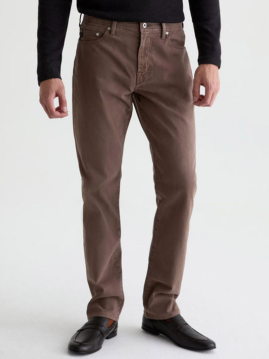 Wearing the AG Jeans Everett Sueded in Sulfur Taupe Grey—brown slim straight jeans made from sueded stretch sateen—a person pairs them with a black long-sleeve shirt and black loafers, standing against a plain background for a subtle vintage look.