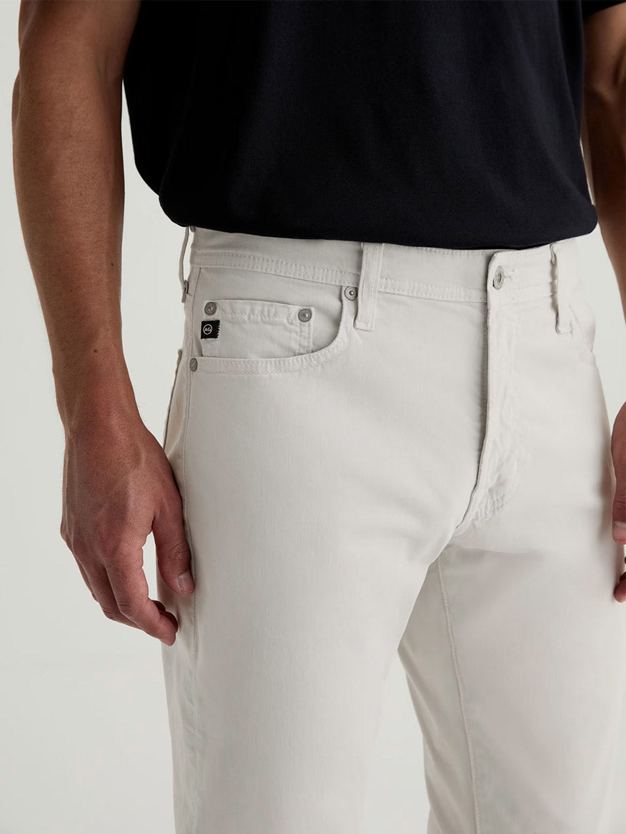 A person wearing AG Jeans Everett Sueded in Smoke, a light grey slim straight jean, paired with a black shirt, shown from waist to mid-thigh with one arm relaxed at their side.