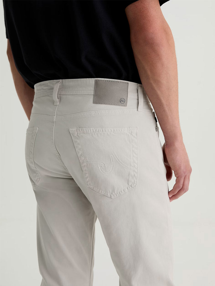 Person wearing AG Jeans Everett Sueded in Smoke, a slim straight style in light beige with visible back pockets and a black shirt, shown from waist down, photographed from behind.