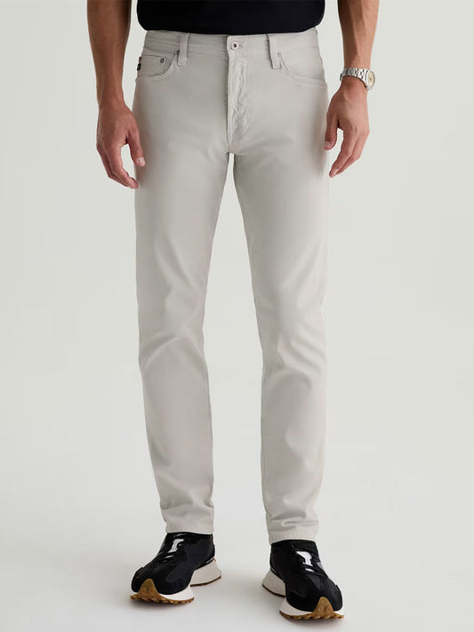 A person wearing AG Jeans Everett Sueded in Smoke, a black shirt, black sneakers with white soles, and a wristwatch stands against a plain background.