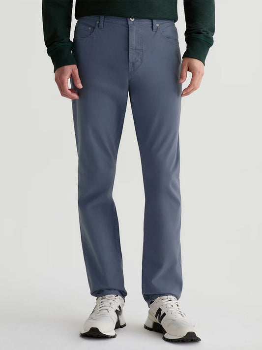 Wearing AG Jeans Everett Sueded in Vintage Cornflower, a dark green sweater, and white New Balance sneakers, a person stands against a plain light background.