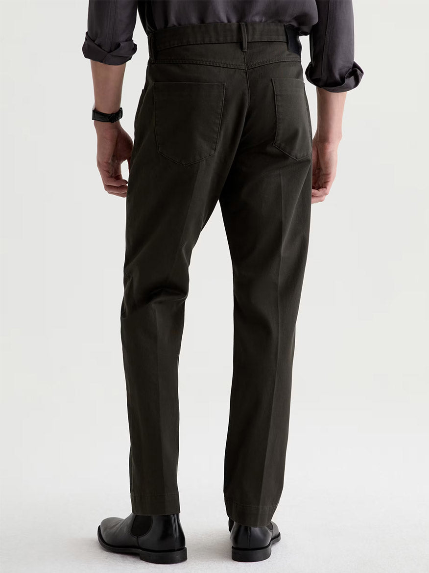 A person stands facing away, wearing AG Jeans Everett Tailored Pant in Forest Ash, a dark long-sleeve shirt, black shoes, and a watch, against a plain light background.
