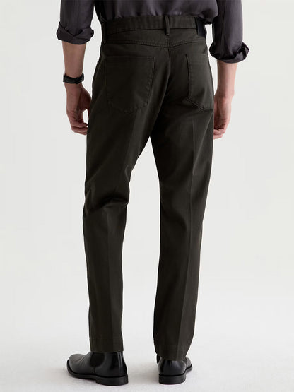 A person stands facing away, wearing AG Jeans Everett Tailored Pant in Forest Ash, a dark long-sleeve shirt, black shoes, and a watch, against a plain light background.
