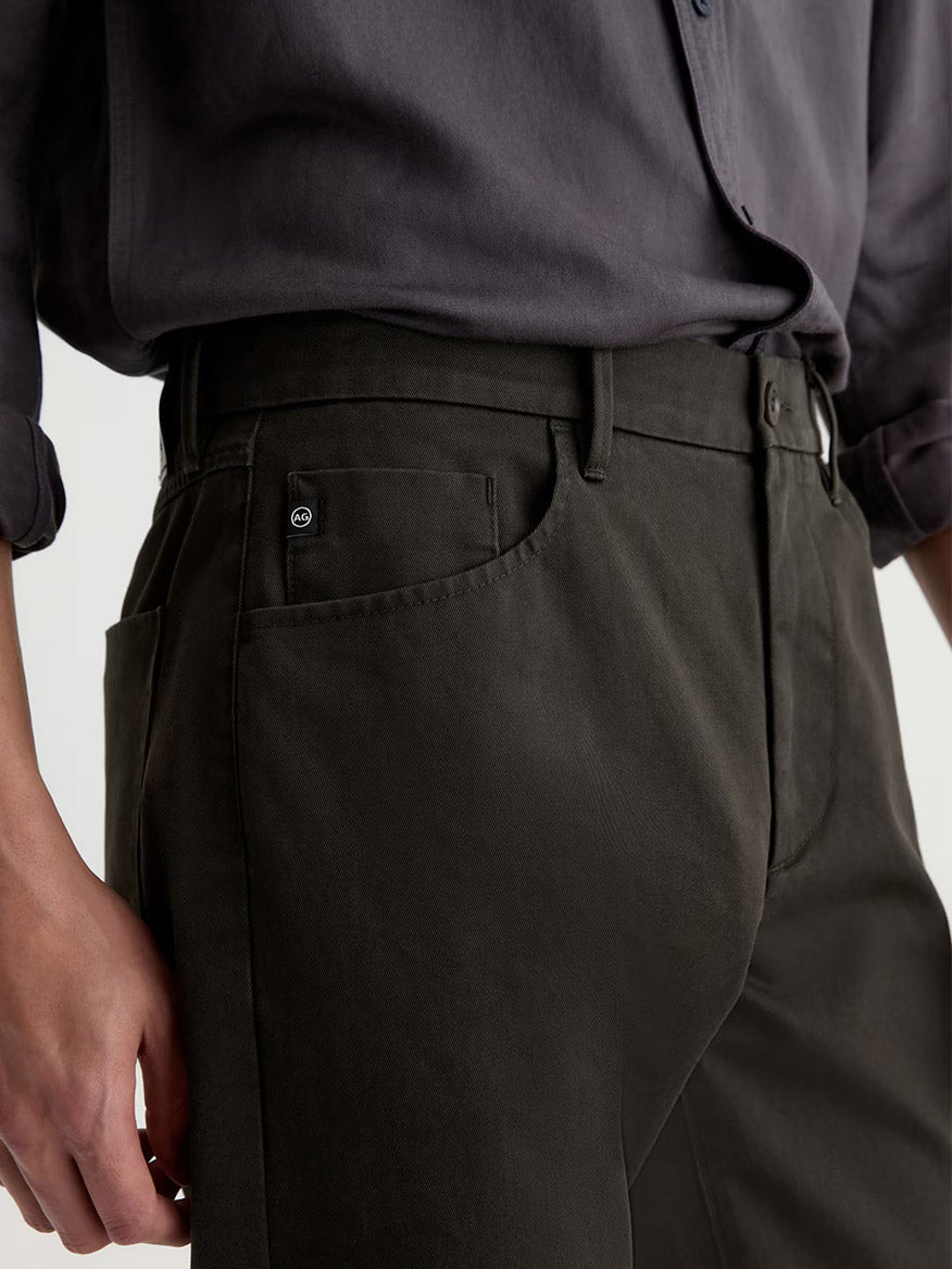 Person wearing AG Jeans Everett Tailored Pant in Forest Ash and a tucked-in dark grey shirt, shown from waist to mid-thigh, with one hand hanging at their side. The slim straight pant style offers a modern silhouette and features a water-resistant finish for added practicality.