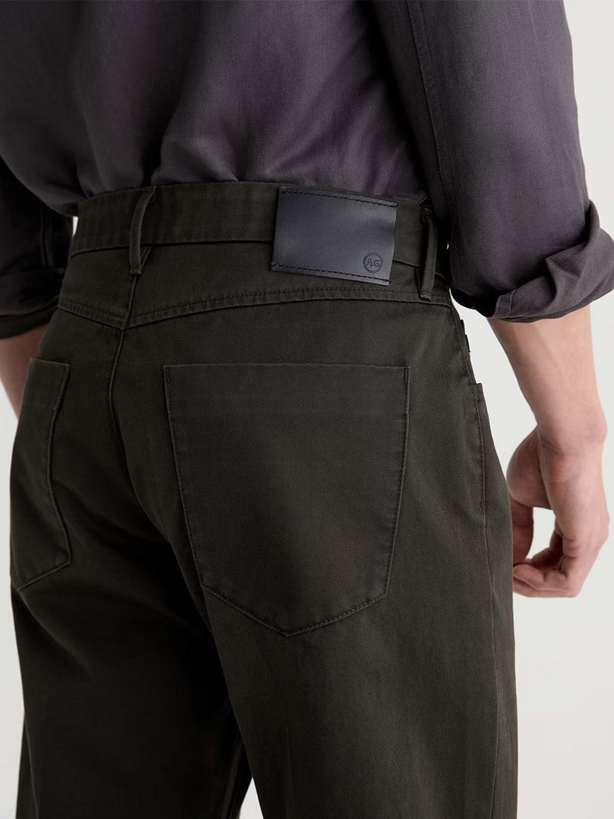 Wearing the AG Jeans Everett Tailored Pant in Forest Ash—slim straight gabardine trousers—with a tucked-in charcoal shirt, and a black leather wallet visible in the back pocket.