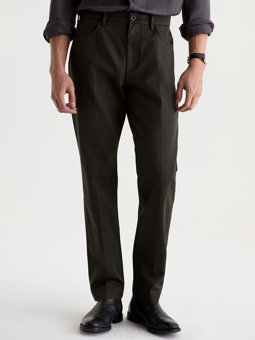 Standing against a plain light background, a person is wearing the AG Jeans Everett Tailored Pant in Forest Ash, styled in a slim straight fit, paired with a dark gray rolled-sleeve shirt and black dress shoes.