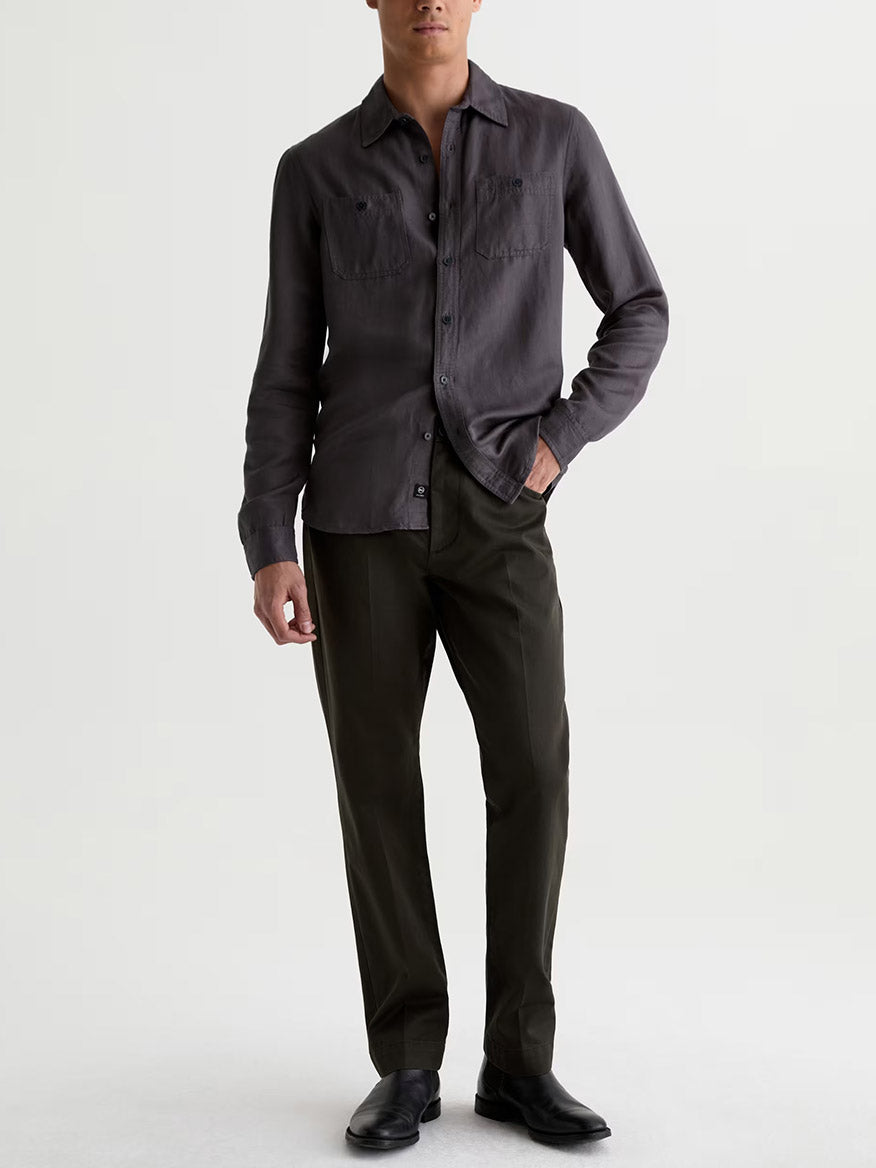 A man stands against a plain background, wearing a dark gray button-up shirt, AG Jeans Everett Tailored Pant in Forest Ash, and black shoes.