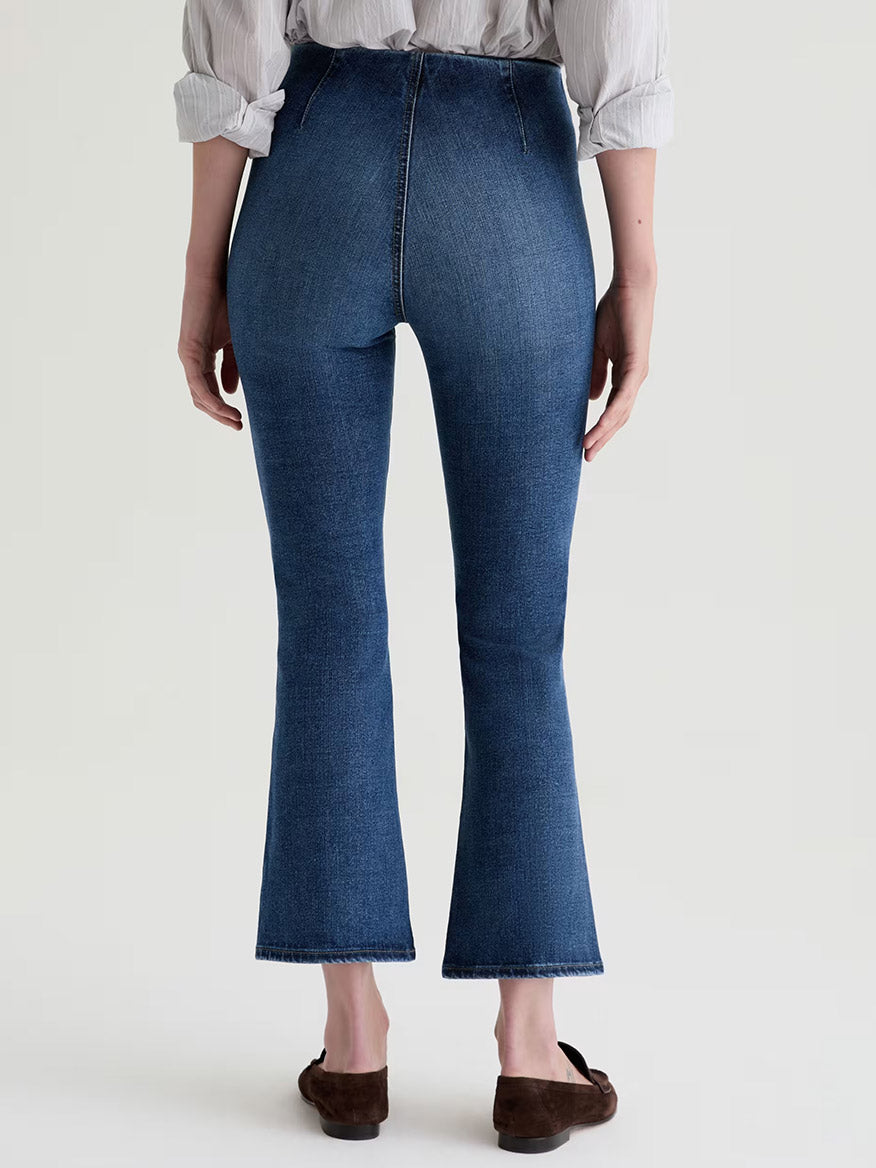 A person is seen from the back wearing AG Jeans Farrah Pull On Mid-Rise Boot Crop in Moonlight, paired with a light shirt and brown flats, against a plain background.