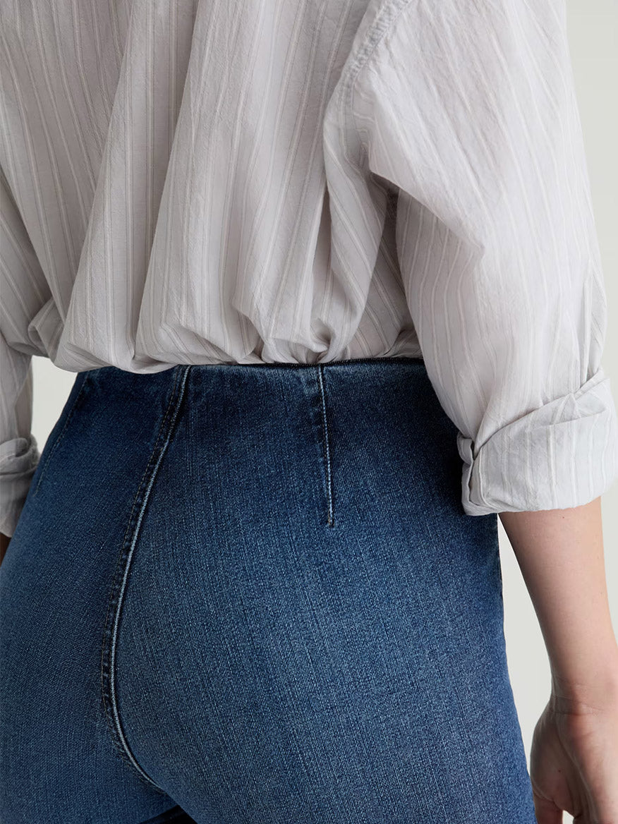 Someone wearing a light-colored, long-sleeve, button-up shirt tucked into AG Jeans Farrah Pull On Mid-Rise Boot Crop in Moonlight, a high-waisted, dark blue jean featuring a flattering bootcut silhouette.