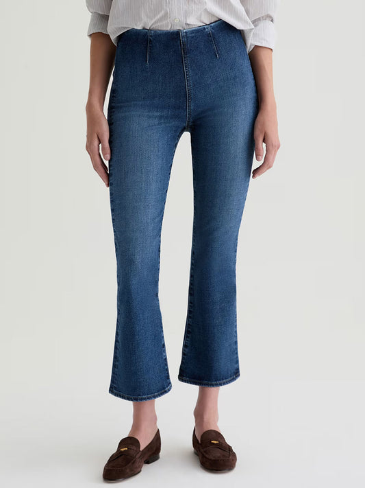 Person wearing AG Jeans Farrah Pull On Mid-Rise Boot Crop in Moonlight with a light-colored, rolled-sleeve shirt and brown loafers, standing against a plain light background.