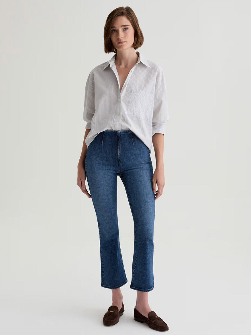 A person stands against a plain background wearing a white button-up shirt, AG Jeans Farrah Pull On Mid-Rise Boot Crop in Moonlight, and brown loafers.