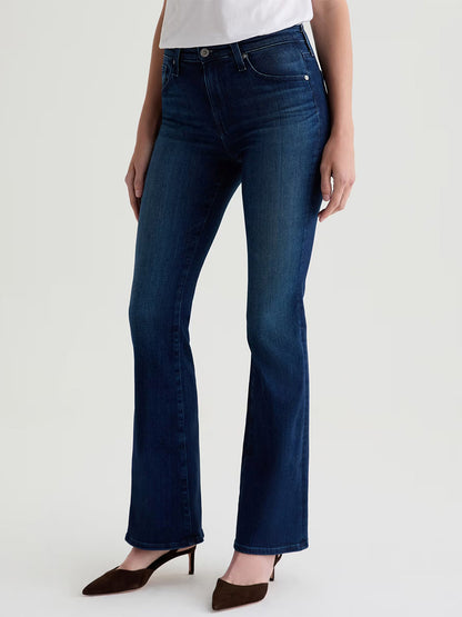 A woman wears AG Jeans Farrah Mid-Rise Boot in Epilogue, paired with a white top and brown pointed-toe heels, standing against a plain light background.