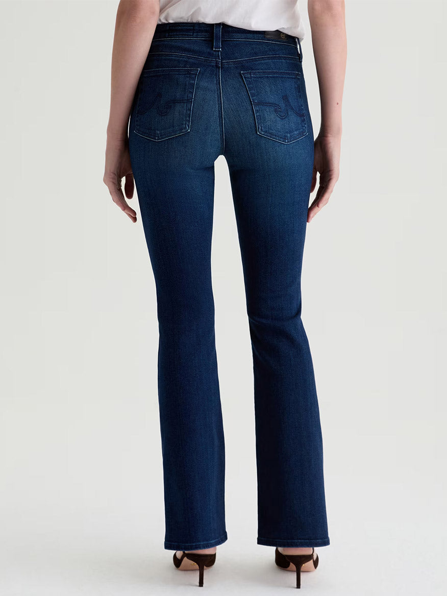 Model in AG Jeans Farrah Mid-Rise Boot in Epilogue, dark blue stretch denim, styled with heeled sandals and shown from the back against a plain light background.