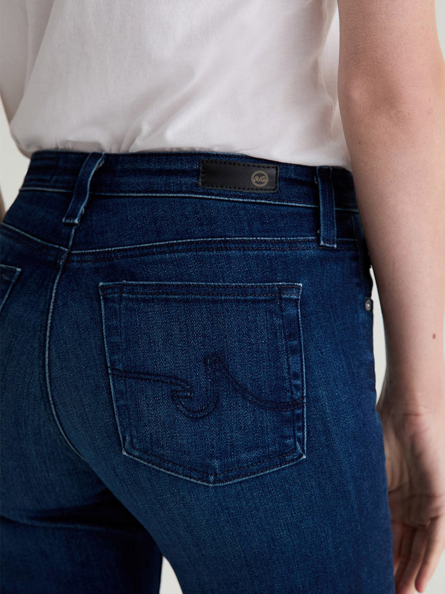 A person is shown from the back wearing AG Jeans Farrah Mid-Rise Boot in Epilogue—dark blue premium Italian denim bootcut jeans with a visible pocket, brand patch, and a white t-shirt.