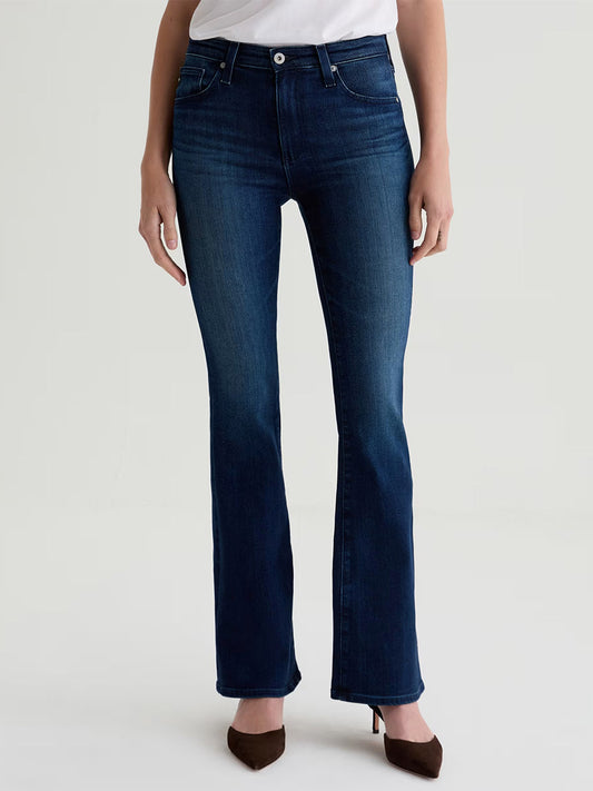 Model wears AG Jeans Farrah Mid-Rise Boot in Epilogue, a dark blue stretch denim, paired with a white top. Only the lower half of her body is visible against a plain light background.
