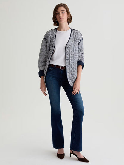 A woman stands against a plain background wearing a patterned jacket, white t-shirt, and the AG Jeans Farrah Mid-Rise Boot in Epilogue, crafted from premium Italian denim, paired with brown heels.