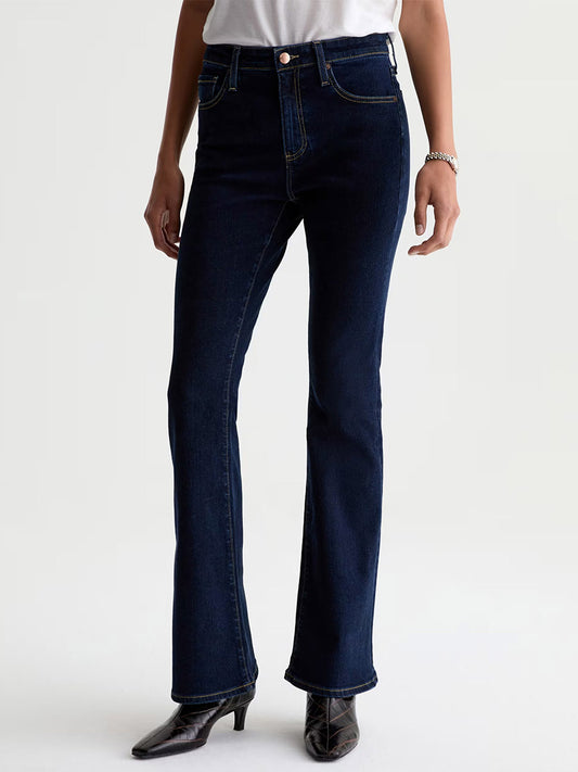 Person wearing a white shirt, AG Jeans Farrah Mid-Rise Boot in Modern Indigo, black pointed boots, and a bracelet, standing against a plain light background.