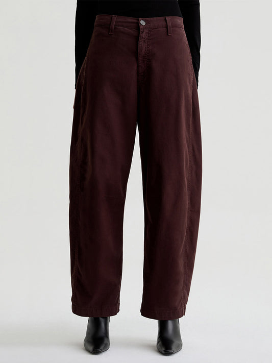 A person wearing AG Jeans Fiona High-Rise Barrel in Coffee Bean, paired with a black top and ankle boots, stands against a plain light background.
