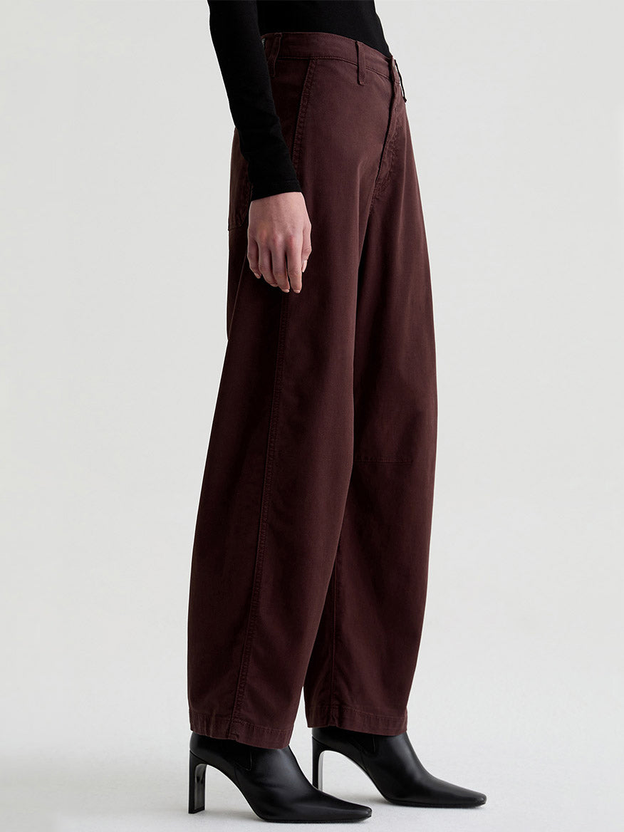 Wearing AG Jeans Fiona High-Rise Barrel in Coffee Bean, a black long-sleeve top, and black heeled boots, a person stands against a plain light background.