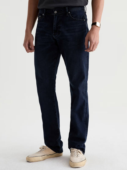 A person wearing a black t-shirt, AG Jeans Graduate in Studio, a wristwatch, and beige shoes stands against a plain light background.