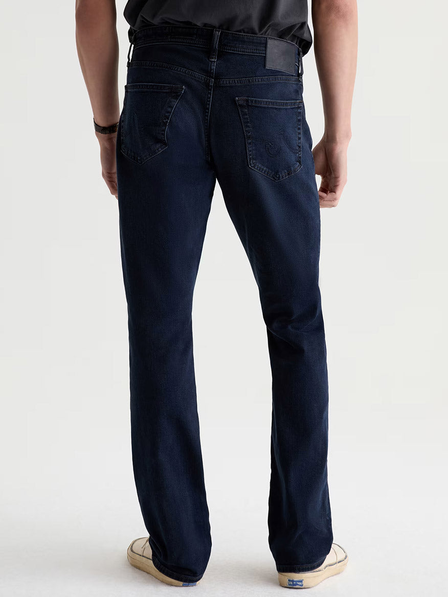 Person wearing AG Jeans Graduate in Studio, a black shirt, and light-colored sneakers, shown from the back against a plain light background.