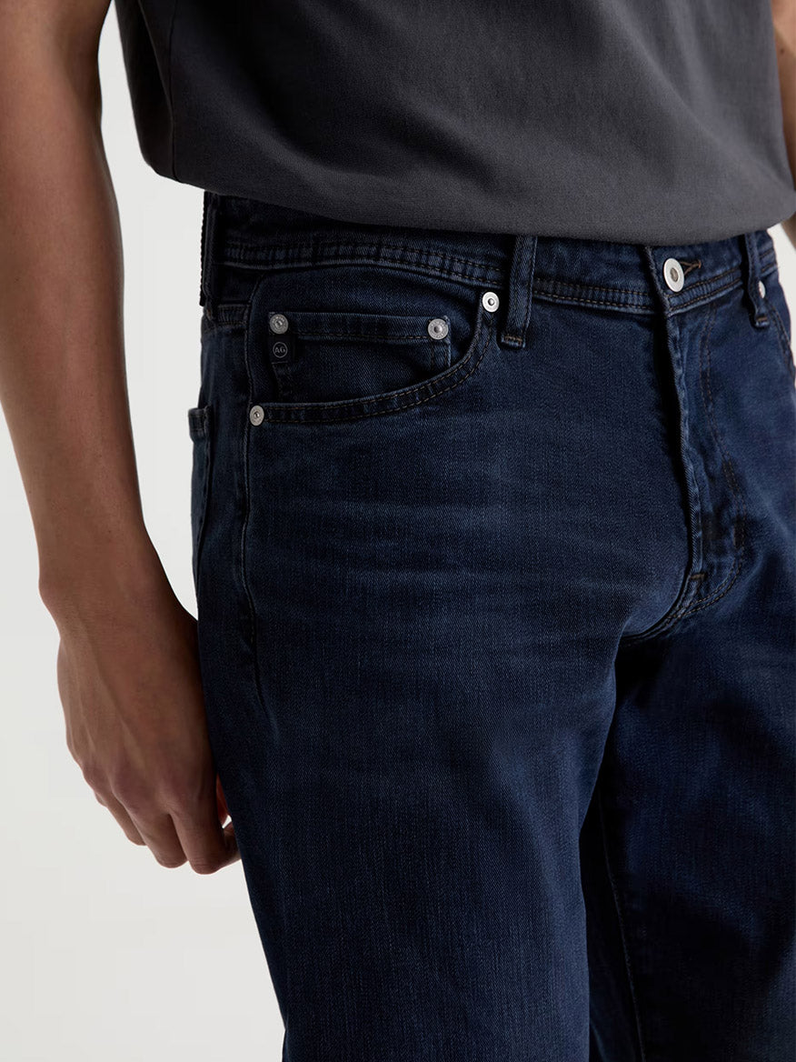 Wearing the AG Jeans Graduate in Studio, a person is shown from the waist to mid-thigh, dressed in dark blue slim straight jeans paired with a dark grey t-shirt, one arm visible at their side.