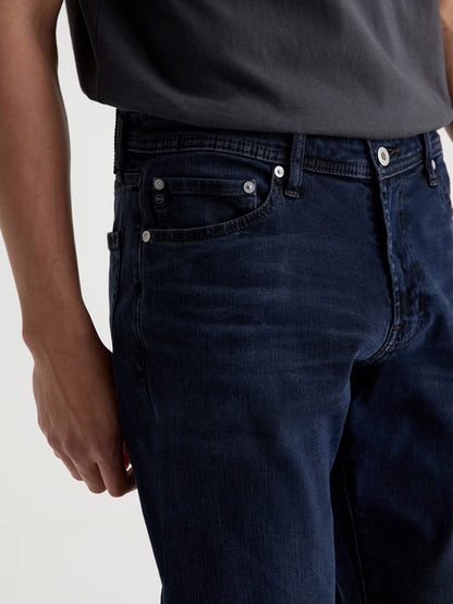Wearing the AG Jeans Graduate in Studio, a person is shown from the waist to mid-thigh, dressed in dark blue slim straight jeans paired with a dark grey t-shirt, one arm visible at their side.